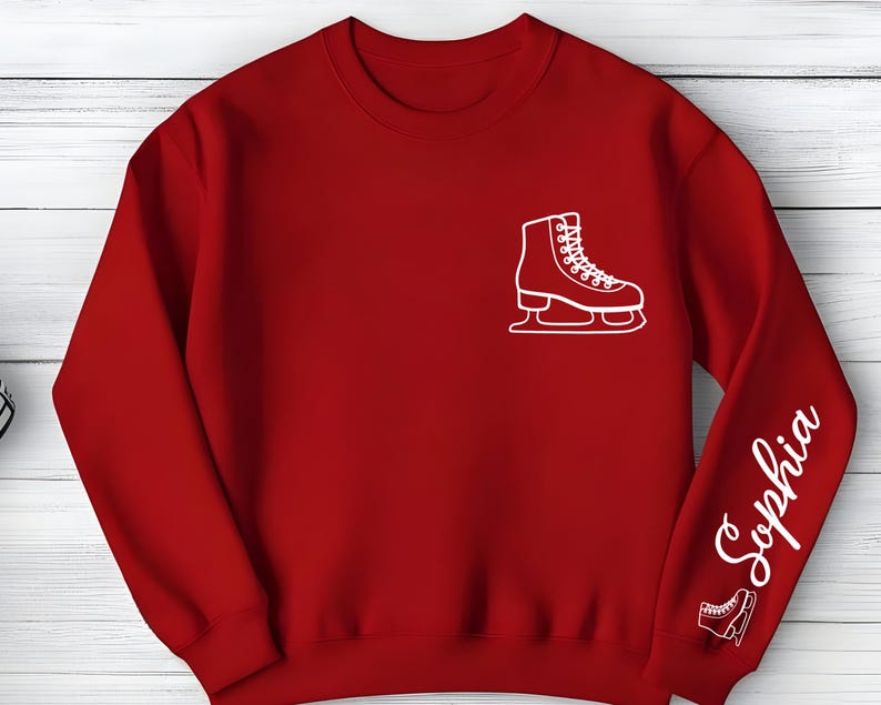 May include: A red sweatshirt featuring a white outline of an ice skate on the chest and the name "Sophia" in white script on the sleeve. A smaller ice skate outline is near the cuff. The sweatshirt is made of a soft material.