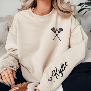 May include: Cream-colored sweatshirt featuring a black lacrosse stick and ball graphic on the chest. The sleeve displays the name "Kylie" in a cursive font, with a small lacrosse stick and ball graphic below the name. The sweatshirt is a casual style.