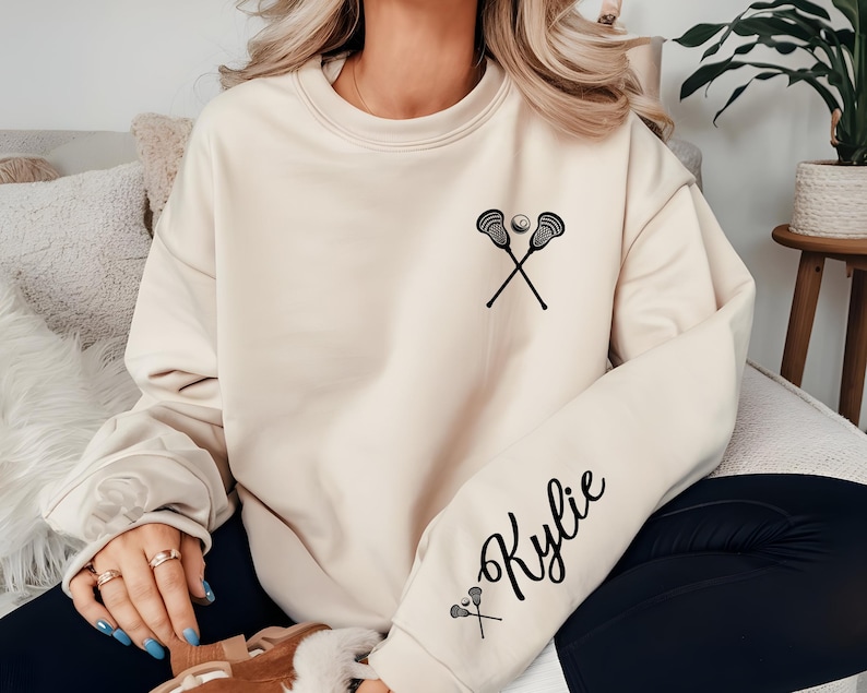 May include: Cream-colored sweatshirt featuring a black lacrosse stick and ball graphic on the chest. The sleeve displays the name "Kylie" in a cursive font, with a small lacrosse stick and ball graphic below the name. The sweatshirt is a casual style.