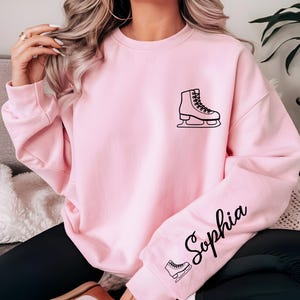 May include: A light pink sweatshirt featuring a black outline of an ice skate on the chest and sleeve. The name "Sophia" is written in cursive on the sleeve. The sweatshirt is a solid color.