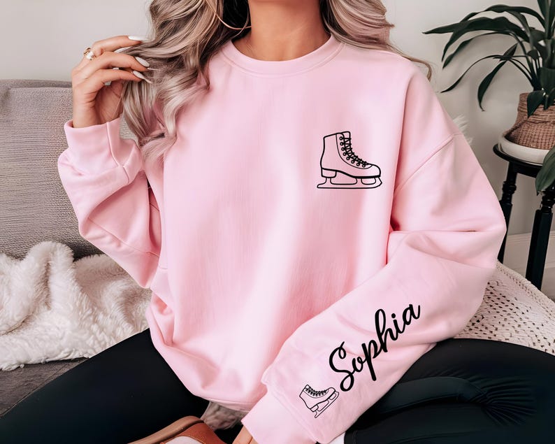 May include: A light pink sweatshirt featuring a black outline of an ice skate on the chest and sleeve. The name "Sophia" is written in cursive on the sleeve. The sweatshirt is a solid color.
