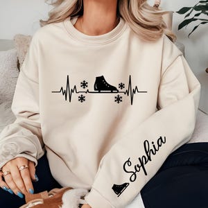 Op de afbeelding: Cr&egrave;mekleurige sweatshirt met een zwarte afbeelding van een schaats ge&iuml;ntegreerd in een hartslaglijn, met sneeuwvlokken. De mouw toont de naam "Sophia" in cursief schrift, plus een klein schaatsicoon. Een gezellig winterthema kledingstuk.