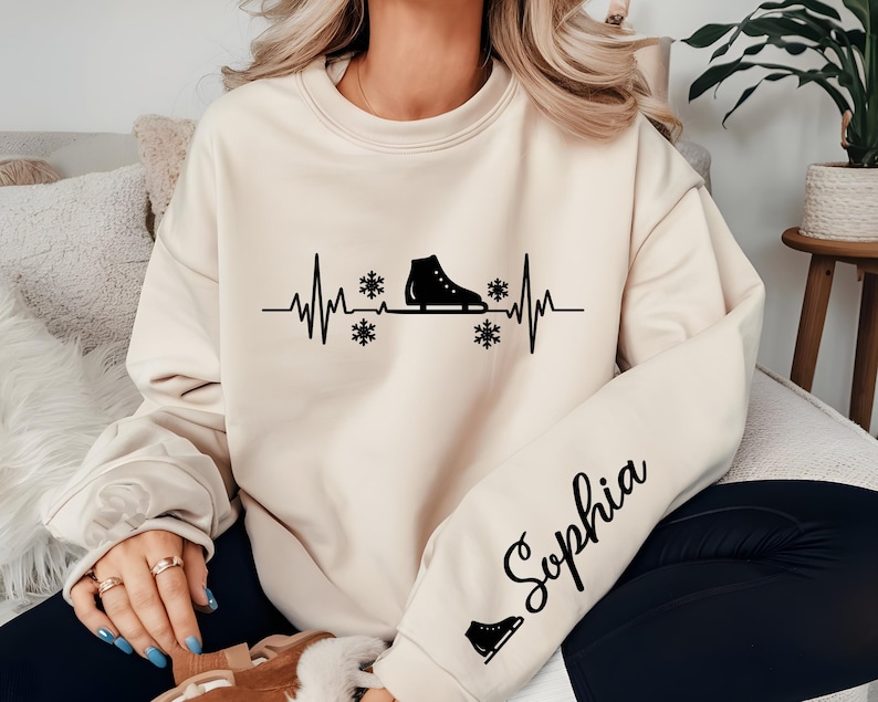 Op de afbeelding: Cr&egrave;mekleurige sweatshirt met een zwarte afbeelding van een schaats ge&iuml;ntegreerd in een hartslaglijn, met sneeuwvlokken. De mouw toont de naam "Sophia" in cursief schrift, plus een klein schaatsicoon. Een gezellig winterthema kledingstuk.
