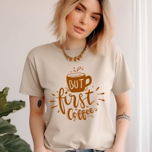 May include: Beige t-shirt with a brown coffee cup graphic and the words "BUT first Coffee" in a stylized font. The shirt is a short-sleeved crew neck style.