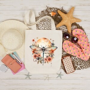 Pu&ograve; includere: Una borsa tote in tela bianca con un designo di libellula. La libellula &egrave; blu e verde con accenti floreali marroni e arancioni. La borsa &egrave; su una superficie di legno bianco con altri oggetti a tema spiaggia.