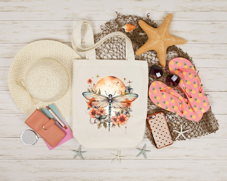 Pu&ograve; includere: Una borsa tote in tela bianca con un designo di libellula. La libellula &egrave; blu e verde con accenti floreali marroni e arancioni. La borsa &egrave; su una superficie di legno bianco con altri oggetti a tema spiaggia.