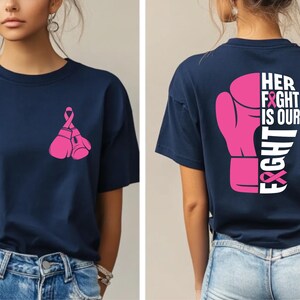 Pode incluir: Camiseta azul marinho com um design de luvas de boxe e fita rosa. A parte de tr&aacute;s da camisa apresenta uma grande luva de boxe rosa com o texto "HER FIGHT IS OUR FIGHT" em branco.
