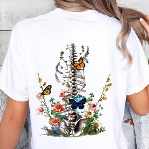 May include: White t-shirt with a graphic of a human spine with flowers and butterflies. The butterflies are orange, blue, and yellow.
