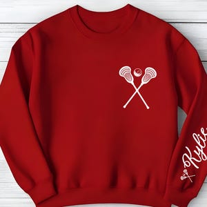 May include: A red sweatshirt featuring a white lacrosse stick and ball design on the chest. The right sleeve displays the name "Kylie" in a cursive font, with a small lacrosse stick graphic below.