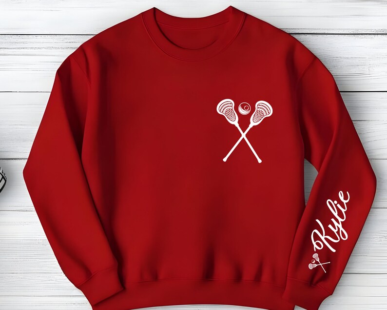 May include: A red sweatshirt featuring a white lacrosse stick and ball design on the chest. The right sleeve displays the name "Kylie" in a cursive font, with a small lacrosse stick graphic below.