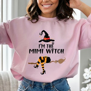 May include: A pink sweatshirt with a graphic of a witch on a broomstick. The witch is wearing a black hat with a red band and orange and black striped stockings. The text on the sweatshirt reads "I'm the Mimi Witch".