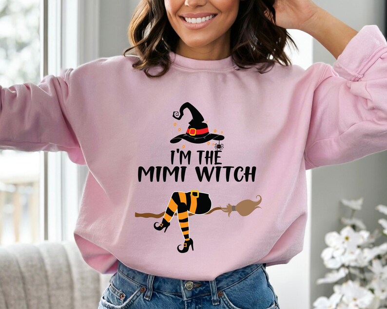 May include: A pink sweatshirt with a graphic of a witch on a broomstick. The witch is wearing a black hat with a red band and orange and black striped stockings. The text on the sweatshirt reads "I'm the Mimi Witch".
