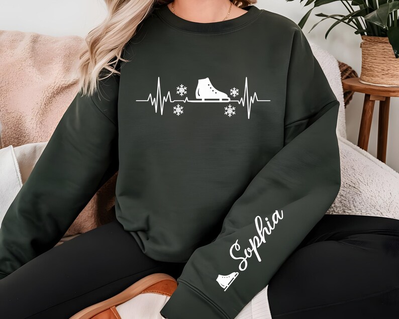 Op de afbeelding: Donkergroene sweatshirt met een witte afbeelding van een schaats, sneeuwvlokken en een hartslaglijn. De mouw heeft de naam "Sophia" in witte letters, plus een klein schaatsicoon. Een comfortabele, casual stijl.