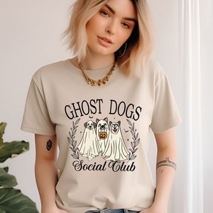 May include: A beige t-shirt with a graphic of three dogs dressed as ghosts. The text "Ghost Dogs Social Club" is printed on the shirt.