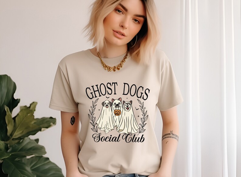 May include: A beige t-shirt with a graphic of three dogs dressed as ghosts. The text "Ghost Dogs Social Club" is printed on the shirt.