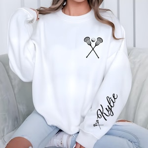 May include: White sweatshirt featuring a black lacrosse stick and ball graphic on the chest. The sleeve has the name "Kylie" in a script font, with a small lacrosse stick graphic. The sweatshirt is a casual, long-sleeved design.