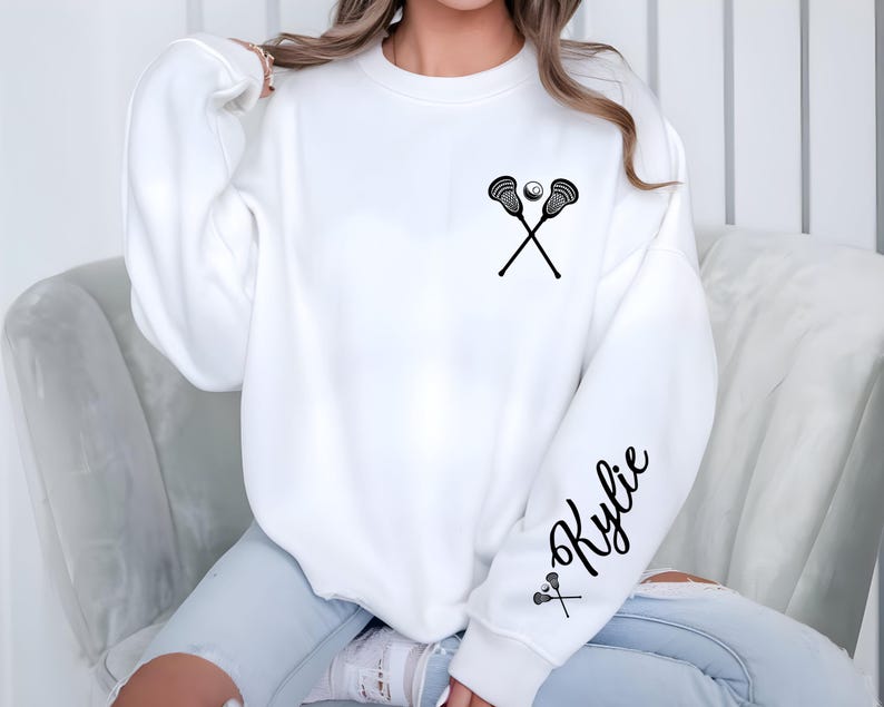 May include: White sweatshirt featuring a black lacrosse stick and ball graphic on the chest. The sleeve has the name "Kylie" in a script font, with a small lacrosse stick graphic. The sweatshirt is a casual, long-sleeved design.