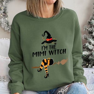 May include: A green sweatshirt with a graphic of a witch on a broomstick. The text on the sweatshirt reads "I'm the Mimi Witch".