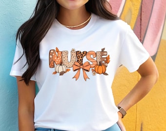 May include: White t-shirt featuring the word "NURSE" in a fall-themed design. The letters have various patterns, including leopard print and fall leaves. A pumpkin, bow, and leaves are also included in the design.