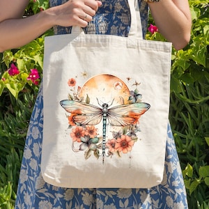 Pu&ograve; includere: Una borsa tote bianca con un design di libellula colorato. La libellula &egrave; circondata da fiori e un cerchio color tramonto. La borsa ha due manici lunghi.