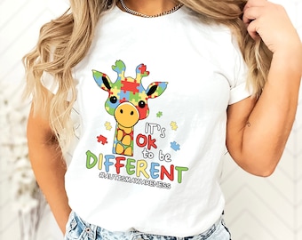 May include: White t-shirt featuring a colorful giraffe with a puzzle-piece design on its head and body. The text "It's ok to be DIFFERENT" is below the giraffe, with the hashtag #AUTISMAWARENESS.