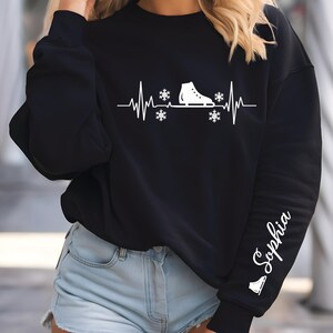 Op de afbeelding: Zwart sweatshirt met een witte afbeelding van een schaats, sneeuwvlokken en een hartslaglijn. De mouw heeft de naam "Sophia" in witte letters. Een winterkledingstuk.
