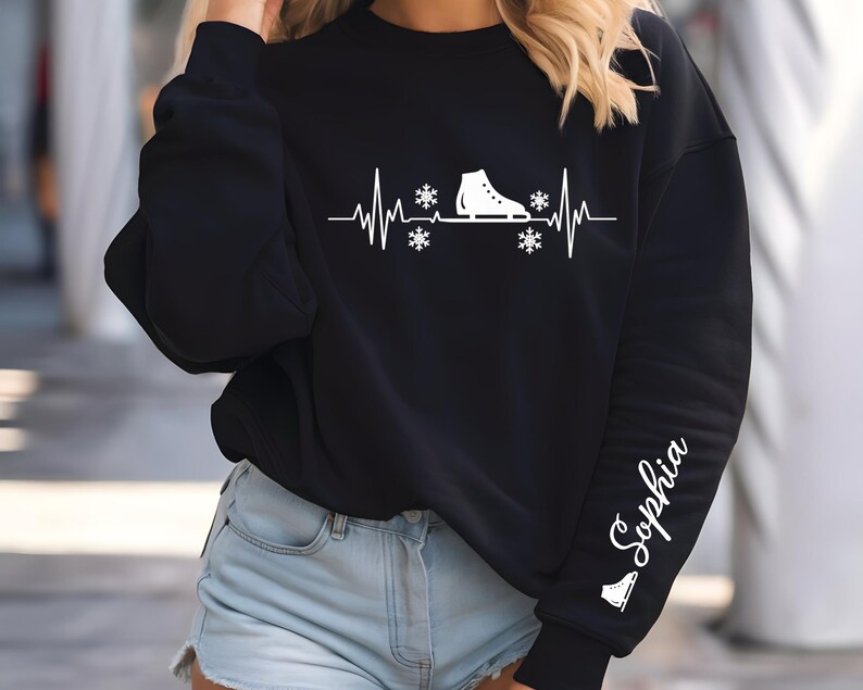 Op de afbeelding: Zwart sweatshirt met een witte afbeelding van een schaats, sneeuwvlokken en een hartslaglijn. De mouw heeft de naam "Sophia" in witte letters. Een winterkledingstuk.