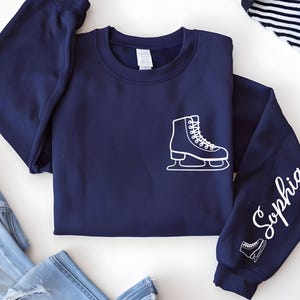 May include: A navy blue sweatshirt with a white graphic of an ice skate on the front and sleeve. The name "Sophia" is written in white script on the right sleeve. A second, smaller ice skate graphic is below the name.