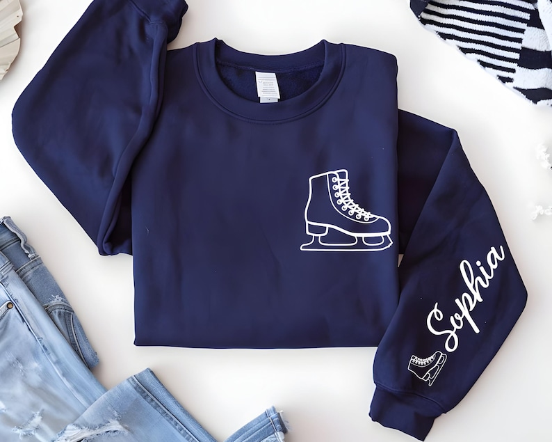 May include: A navy blue sweatshirt with a white graphic of an ice skate on the front and sleeve. The name "Sophia" is written in white script on the right sleeve. A second, smaller ice skate graphic is below the name.
