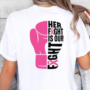 Pode incluir: Camiseta branca com um desenho de luva de boxe rosa. O texto "HER FIGHT IS OUR FIGHT" &eacute; impresso em preto e rosa, com fitas rosas incorporadas no design. A camiseta &eacute; de estilo casual.