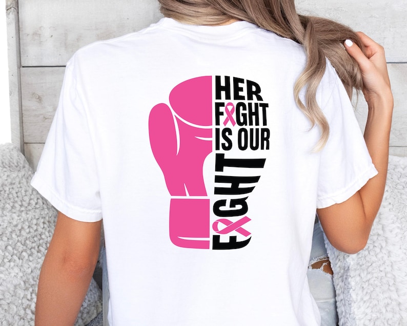Pode incluir: Camiseta branca com um desenho de luva de boxe rosa. O texto "HER FIGHT IS OUR FIGHT" &eacute; impresso em preto e rosa, com fitas rosas incorporadas no design. A camiseta &eacute; de estilo casual.