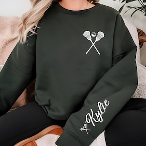 May include: Dark green sweatshirt featuring a white lacrosse stick and ball graphic on the chest. The sleeve displays the name "Kylie" in white script, along with a small lacrosse stick graphic. The sweatshirt is a casual, comfortable style.