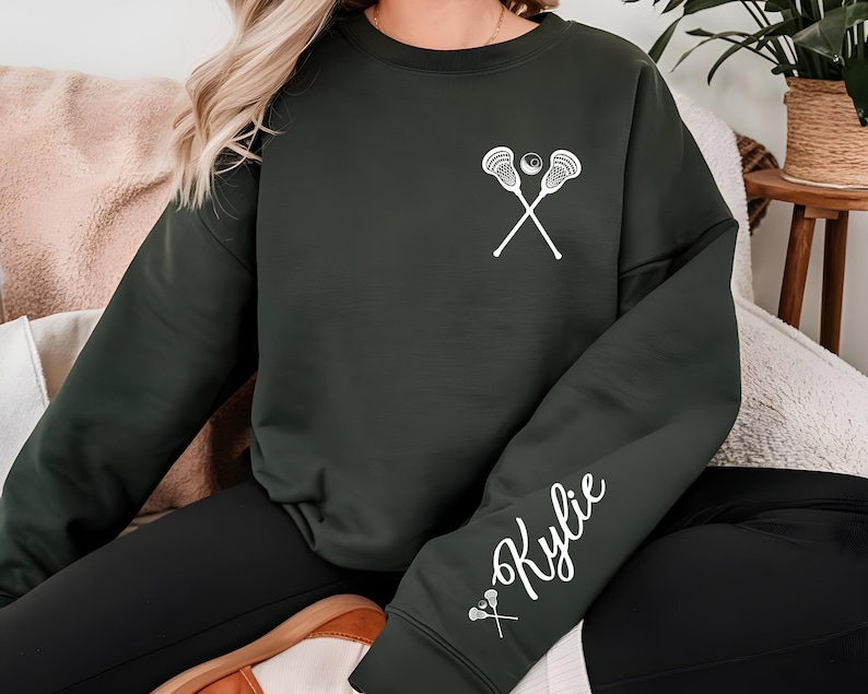 May include: Dark green sweatshirt featuring a white lacrosse stick and ball graphic on the chest. The sleeve displays the name "Kylie" in white script, along with a small lacrosse stick graphic. The sweatshirt is a casual, comfortable style.