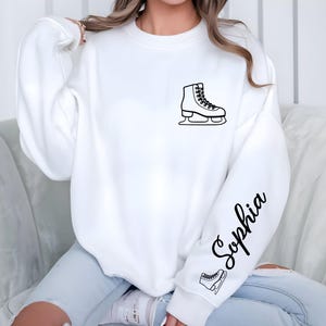 May include: White sweatshirt featuring a black outline of an ice skate on the chest and sleeve. The name "Sophia" is written in cursive on the sleeve. A small ice skate graphic is below the name.