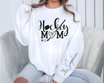 May include: White sweatshirt with the text "Hockey Mom" in black script, featuring a hockey puck, stick, and heart design. The sleeve has the name "X. Sophia" in cursive. A comfortable, casual top for hockey fans.