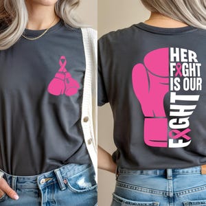 Pode incluir: Camiseta cinza escuro com um design de luva de boxe e fita rosa. A frente apresenta duas luvas e uma fita. A parte de tr&aacute;s exibe uma luva grande com o texto "HER FIGHT IS OUR FIGHT".