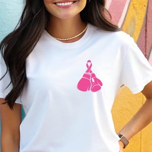 Pode incluir: Camiseta branca com um la&ccedil;o rosa e luvas de boxe, um s&iacute;mbolo de conscientiza&ccedil;&atilde;o sobre o c&acirc;ncer de mama. A camiseta &eacute; feita de um material macio e confort&aacute;vel.