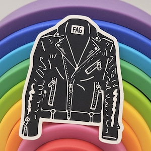 May include: A black and white sticker of a leather jacket with the word "FAG" on the collar. The sticker is placed on top of a wooden rainbow arch toy with red, pink, orange, yellow, green, blue, and purple arches.