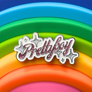 Op de afbeelding: Een witte emaille pin met de woorden "Prettyboy" in roze glitterletters. De pin is omringd door zilveren sterren en is geplaatst tegen een regenboogachtergrond.