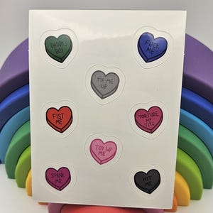 May include: A sheet of heart-shaped stickers with various phrases, including "Daddy's Boy," "Fist Me," and "Spank Me." The stickers are in different colors, such as green, red, and pink. The sheet is resting on a wooden rainbow.