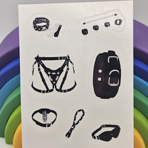 May include: A sheet of black and white stickers featuring various BDSM accessories. The stickers include a studded collar with a chain, a harness, a blindfold, a paddle, a gag, and a wrist restraint.