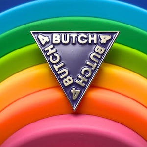 May include: A purple triangle-shaped enamel pin with the text "4 BUTCH 4" written around the perimeter in white lettering.