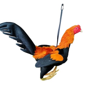 Può includere: Un giocattolo di peluche a forma di gallo nero e arancione con cresta e bargigli rossi. Il gallo ha una coda e ali nere ed è attaccato a uno spago nero.