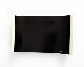 May include: A rectangular black sticker with a glossy finish and white peel-away backing strips on the left and right edges. The text SLAP ADDICT STICKER CO is printed in small white letters in the bottom right corner of the black surface.
