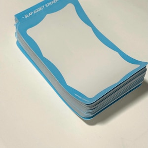 May include: A stack of rectangular stickers with rounded, scalloped edges and a light blue border surrounding a white center. The text SLAP ADDICT STICKER CO. is printed in white on the top blue border of each sticker in the stack.