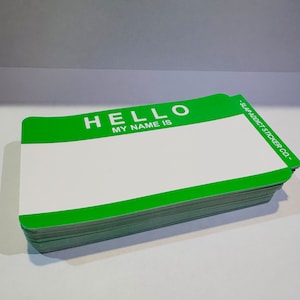 May include: A stack of green and white name tags. The top tag reads "HELLO MY NAME IS" in white text against a green background. The bottom right corner of the tags reads "- SLAP ADDICT STICKER CO." in white text.
