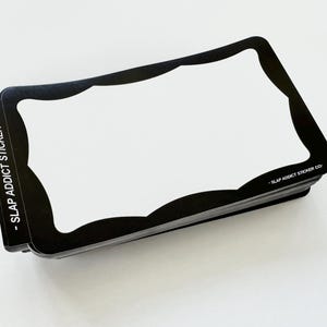Puede incluir: Una pila de pegatinas rectangulares con un borde negro y un centro blanco. El borde negro presenta un borde interior festoneado. El texto SLAP ADDICT STICKER CO. está impreso en blanco en el lado izquierdo y en la esquina inferior derecha.