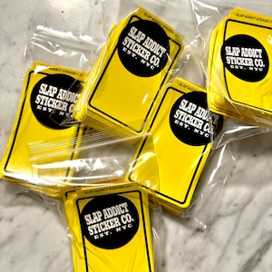 May include: Multiple packets of yellow stickers with black text and a black circle logo. The text reads "SLAP ADDICT STICKER CO. EST. NYC". The stickers are rectangular with rounded corners and are packaged in clear plastic bags.