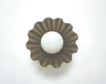 May include: A modern, decorative wall sconce featuring a white globe light centered within a brown, scalloped metal shade. The shade has a textured, pleated design. The fixture is mounted on a white wall.