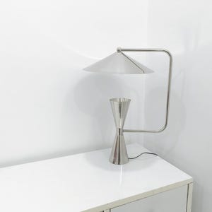 May include: A contemporary desk lamp in a silver finish, featuring a conical lampshade and an hourglass-shaped base. The lamp has a curved arm and is positioned on a white surface. The design is minimalist and modern.